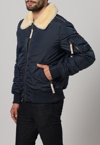 Marinblå bomberjacka med en mjuk krämfärgad fuskpälskrage, ribbade manschetter och fåll, dragkedjefickor samt texturerade detaljer på ärmarna.