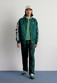 Vihreä pommitakki valkoisilla raidoilla, kerrostettuna yhteensopivan hupparin ja housujen päälle. Valkoinen baseball-lippis ja vihreät lenkkarit viimeistelevät asun.