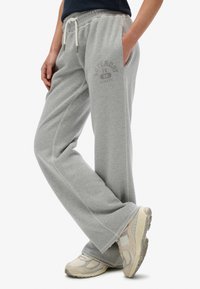 Persoon in grijze sportieve wijde sweatpants met het logo "Superdry 1986 Athletic" en beige New Balance sneakers, hand in de zak.
