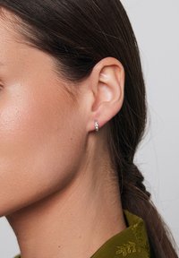 Srebrne kolczyki w kształcie kół z małymi, fasetowanymi akcentami, pokazane noszone w uchu osoby z prostymi ciemnymi włosami i oliwkowo-zielonym strojem.