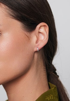 Srebrne kolczyki w kształcie kół z małymi, fasetowanymi akcentami, pokazane noszone w uchu osoby z prostymi ciemnymi włosami i oliwkowo-zielonym strojem.