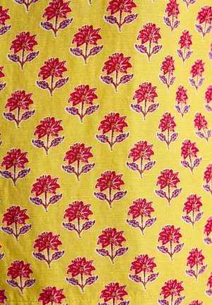 Keltainen kangas, jossa on toistuva kuvio punaisista kukista vihreillä lehdillä. Suunnittelussa on yksityiskohtaisia kukkakoristeita ja teksturoitu pinta.