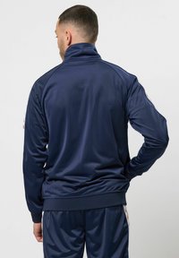 Jachetă de trening bleumarin cu guler înalt, textură netedă și manșete adaptate. Include accente în dungi contrastante pe mâneci.