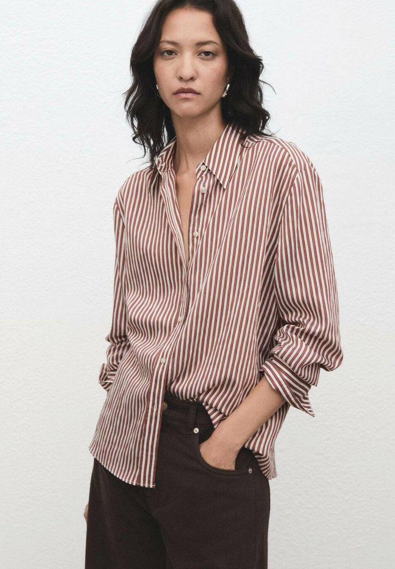Striped shirt in burgundy and white, button-up style with a collar and rolled sleeves. Worn over dark trousers, featuring a relaxed fit.