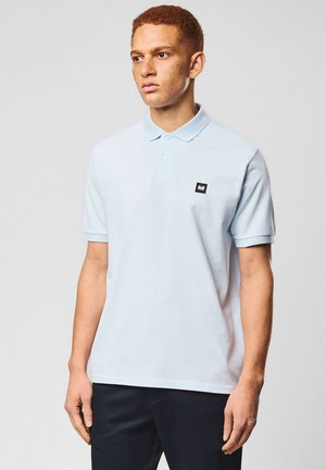 Giovane uomo con capelli rossi corti che indossa una polo azzurra, in piedi davanti a uno sfondo grigio uniforme.