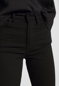 Primer plano de una persona vestida con jeans negros ajustados y una blusa negra metida por dentro, contra un fondo neutro.