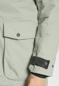 Veste matelassée vert clair avec une poche à bouton-pression et une sangle de poignet noire réglable ; texture lisse avec détails de couture.