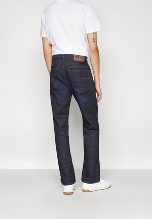 Personne portant un jean bleu foncé, un t-shirt blanc et des baskets blanches, debout de dos sur un fond uni clair.