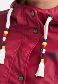 Red waterproof jacket with a high collar, white drawstrings featuring colorful beads, and a black umbrella logo on the front.
