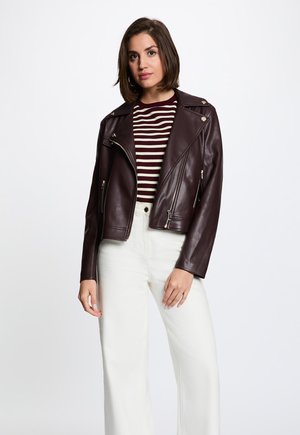 Donna con giacca di pelle marrone scuro sopra una camicia a righe bordeaux e bianca con pantaloni bianchi a vita alta, in piedi su uno sfondo neutro.
