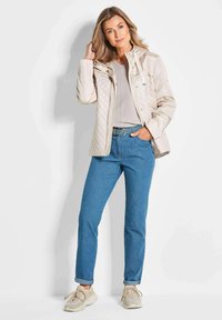 Beige gesteppte Jacke mit Kapuze, kombiniert mit einem hellgrauen Oberteil und blauen Jeans. Der Look wird mit beigen Schuhen abgerundet.