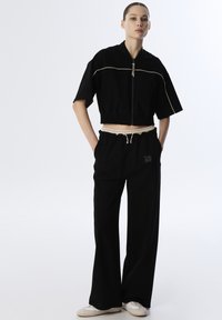 Black cropped jacket with a zip closure and cream piping, paired with black wide-leg pants featuring a drawstring waist and logo detail.