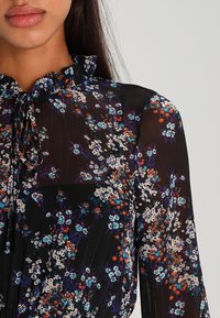 Musta läpinäkyvä paita, jossa on sinisiä, valkoisia ja oransseja kukkakuoseja. Varustettu röyhelökauluksella ja nauhalla kaulalla.