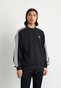 Svart sweatshirt med vitt tre-stripesdesign på ärmarna, rund hals, lös passform och liten logotyp på bröstet. Slät tygtextur.