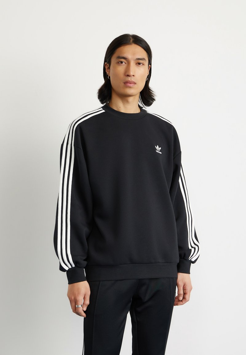 Svart sweatshirt med vitt tre-stripesdesign på ärmarna, rund hals, lös passform och liten logotyp på bröstet. Slät tygtextur.