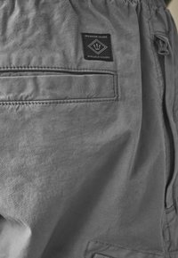 Șorturi gri din material textil, dotate cu un buzunar cu fermoar și eticheta care citește "Produse Durabile de Calitate Premium". Design neted, casual, cu textură ușoară.