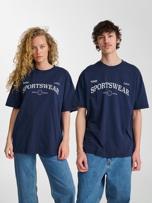 To unge voksne iført marineblå "SPORTSWEAR" t-shirts og blå jeans står side om side mod en ensfarvet hvid baggrund.