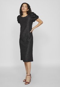 Μαύρο φόρεμα με υφή υφάσματος, κοντά φουσκωτά μανίκια και μήκος μέχρι το γόνατο. Φορεμένο με ψηλοτάκουνα σανδάλια.