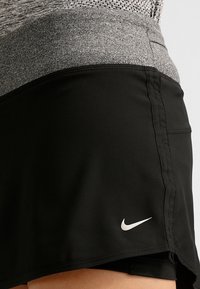 Falda atlética negra con una cintura de un gris jaspeado. Presenta un logo de Nike en la parte inferior izquierda. La tela parece suave con una textura ligera.