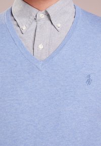 Pull bleu clair en maille à col en V superposé sur une chemise grise à boutons, avec des boutons blancs et un petit logo brodé.