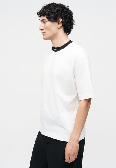 Νέος άνδρας με σγουρά μαλλιά που φοράει λευκό κοντομάνικο πουκάμισο με μαύρη λεπτομέρεια γιακά και μαύρο παντελόνι, στέκεται στο προφίλ με λευκό φόντο.