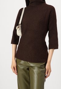 Pull en maille marron à col montant, manches courtes et finition texturée, associé à un pantalon en cuir vert olive et un petit sac blanc.