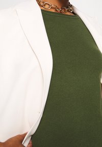 Vit blazer över en grön ribbad topp med rund halsringning. Ett kraftigt guldhalsband ger detaljer till halsringningen.