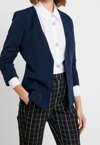 Blazer blu navy con un design aderente, maniche arricciate e polsini bianchi. Abbinato a una camicia bianca con bottoni decorativi e pantaloni a quadri.