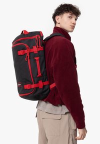 Czarna plecak z czerwonymi akcentami, z regulowanymi paskami, wieloma kieszeniami i trwałym wykończeniem z tkaniny. Noszony na plecach modela.