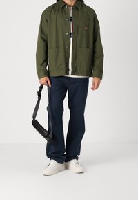 Olive zelená bavlněná bunda s zapínáním na knoflíky, našitým kapsou a dvěma předními kapsami, doplněná o tmavě modré kalhoty a bílé tenisky.