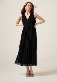 Zwarte geplooide maxi-jurk met een v-hals en mouwloos ontwerp. Heeft een textuur in het lijfje en een vloeiende rok. Afgemaakt met zilveren banden sandalen.