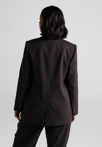 Zwarte op maat gemaakte blazer met gestructureerde schouders, een ventilerende achterkant en vierknopenmanchetten. Gladde stof met een matte afwerking.