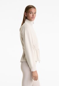 Svetlo béžová bunda na zips s vysokým golierom, sčereným pásom a hladkým materiálom. Obsahuje dlhé rukávy a voľný strih.