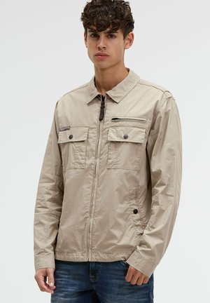 Junger Mann trägt eine beige Jacke mit Reißverschluss und zwei Klappentaschen auf der Brust sowie dunkelblaue Jeans, steht vor einem schlichten Hintergrund.