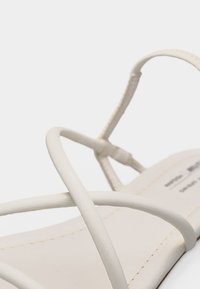 Κοντινό πλάνο ενός λευκού σανδάλιου με λεπτούς, διασταυρούμενους ιμάντες και μινιμαλιστικό σχέδιο σε λευκό φόντο.