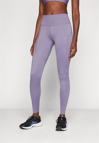Lila sport leggings magas derékkal, oldalsó zsebekkel és sima, rugalmas anyagból. Fekete sportcipővel párosítva.