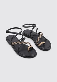 Sandalias de cuero negro con tiras finas y un acento trenzado en tono marrón cruzando el dedo del pie. Presenta una suela plana y una correa en el tobillo ajustable.
