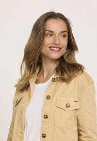 Chemise khaki à boutons avec un col, deux poches poitrine et détails de boutons marron sur un t-shirt blanc. Texture de tissu lisse.