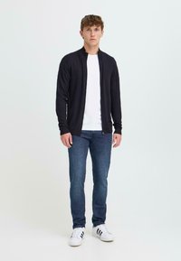 Crna zip-up cardigan preko bijele majice, uparen s tamnim trapericama i bijelim tenisicama sa crnim prugama. Jednostavan, prilagođen dizajn.