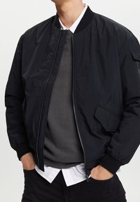 Homme portant une veste bomber noire sur un pull gris et une chemise blanche, les mains dans les poches, debout devant un fond blanc.