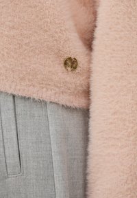 Pull en laine rose clair avec un bouton circulaire doré, associé à un tissu gris texturé, avec des plis visibles et une finition lisse.