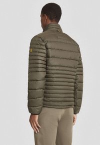 Olijfgroene pufferjack met horizontaal gestikte secties, hoge kraag en een klein logo-opdruk op de linkermouw. Gladde, glanzende textuur.