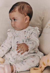 Body de bebé blanco con textura acanalada, estampado floral, hombros con volantes y borde delicado. Niño sentado sobre una tela de color claro cerca de juguetes de madera.