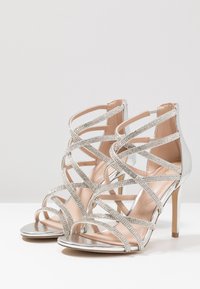 Verspielte High-Heelsandalen aus silbernem, metallic-farbenem Material, ausgestattet mit sich kreuzenden Riemen, die mit Strasssteinen verziert sind, und einem schlanken Absatz.