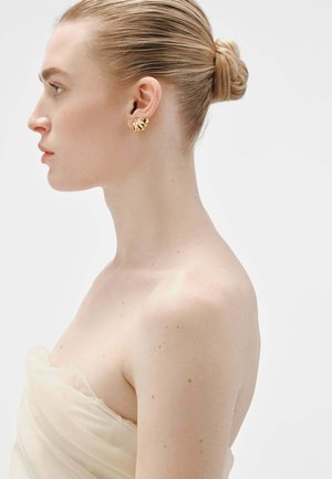 Pendientes en forma de hoja de oro en un modelo, con un acabado pulido y un diseño texturizado. El modelo lleva una blusa de tela ligera y drapeada.