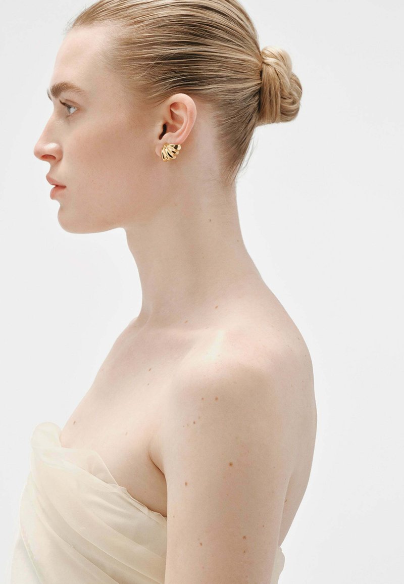Pendientes en forma de hoja de oro en un modelo, con un acabado pulido y un diseño texturizado. El modelo lleva una blusa de tela ligera y drapeada.