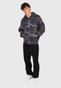 Ung mand iført en sort og grå camouflage zip-up hættetrøje, sorte bukser og hvide sneakers, stående med hænderne i lommerne.