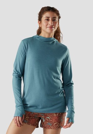 Vrouw met krullend haar, gekleed in een teal gekleurde hoodie met lange mouwen en bruine korte broek met patroon, staand tegen een effen lichte achtergrond.