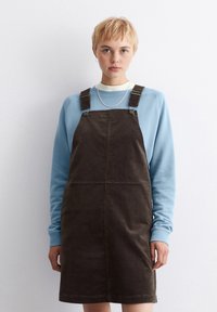 Braunes Corduroy-Overall-Kleid mit verstellbaren Trägern über einem hellblauen Sweatshirt, das über einen runden Halsausschnitt und eine dezente gerippte Textur verfügt.