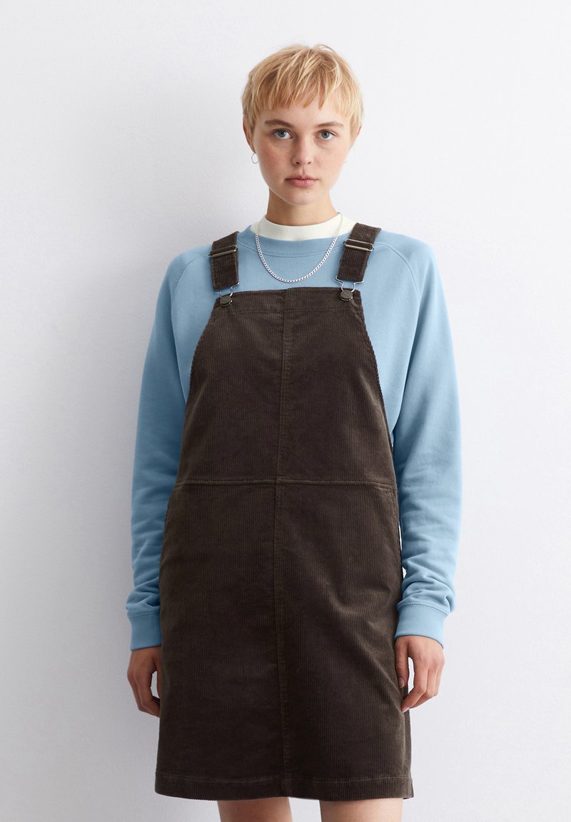Braunes Corduroy-Overall-Kleid mit verstellbaren Trägern über einem hellblauen Sweatshirt, das über einen runden Halsausschnitt und eine dezente gerippte Textur verfügt.